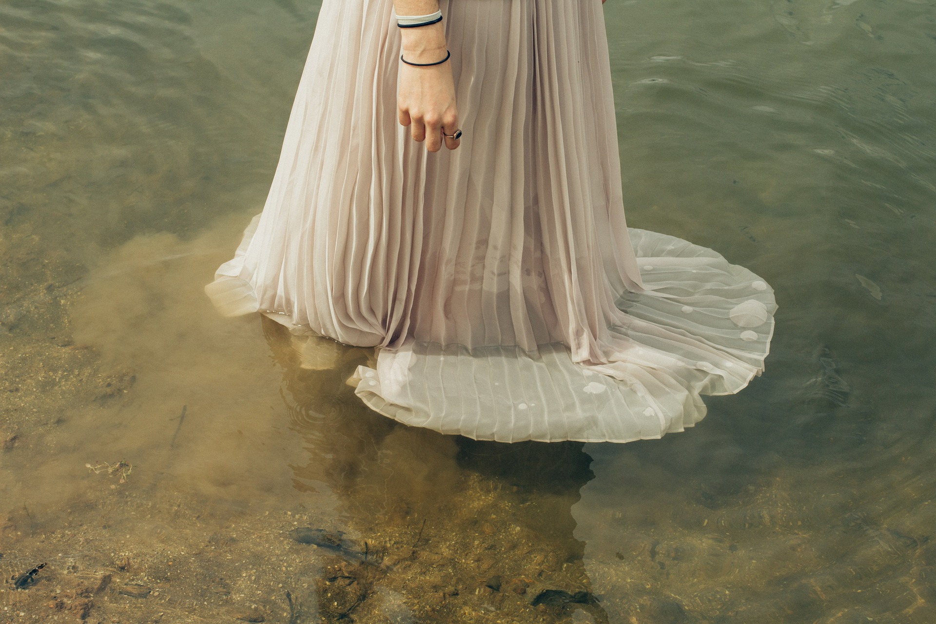 Bottom half of a woman standing in a shallow body of water. Her white skirt spreads out in the water